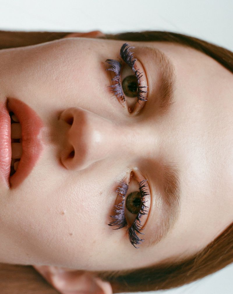 Close-up portrait of a woman's face with lilac-colored mascara on her eyelashes, fair skin, and light brown hair against a white background.