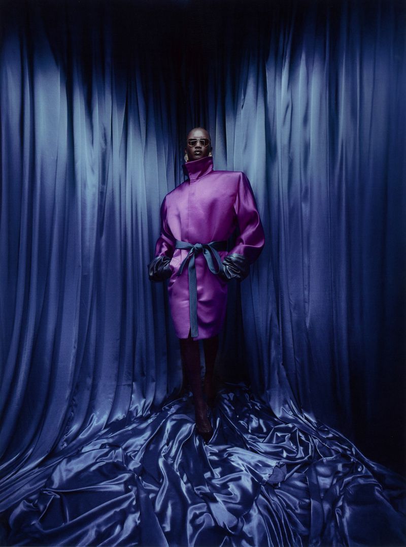 Woman with short hair poses in purple dress with blue silk background and train, runway fashion.