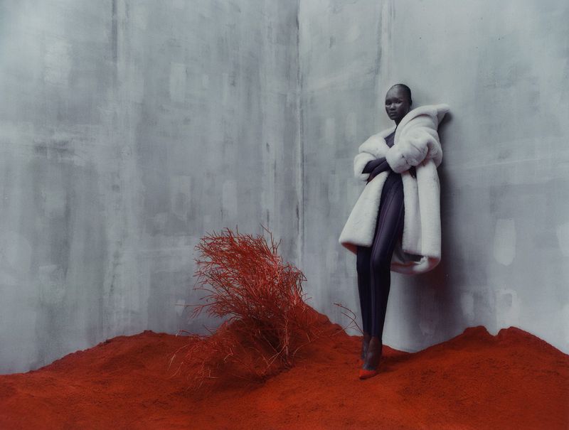 A South Sudanese woman wearing a voluminous white fur coat, standing on red earth in front of a monochromatic gray wall in a studio.