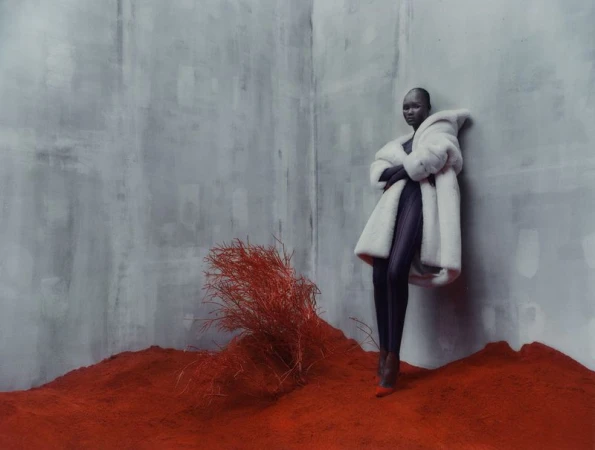 A South Sudanese woman wearing a voluminous white fur coat, standing on red earth in front of a monochromatic gray wall in a studio.
