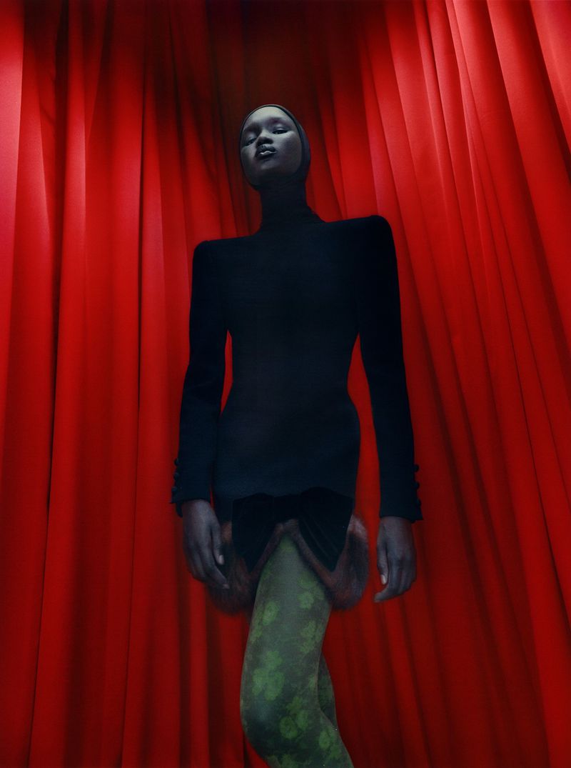 Fashion shot of a slender Black woman posing in a black top and green patterned tights against a backdrop of red curtains.