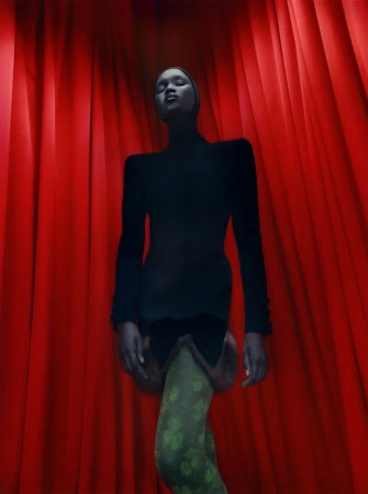 Fashion shot of a slender Black woman posing in a black top and green patterned tights against a backdrop of red curtains.