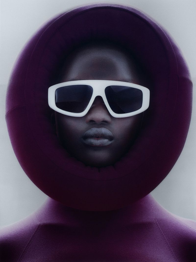 Fashion portrait of an adult woman of color wearing white sunglasses and a plum-colored garment with a circular hood against a light grey background.