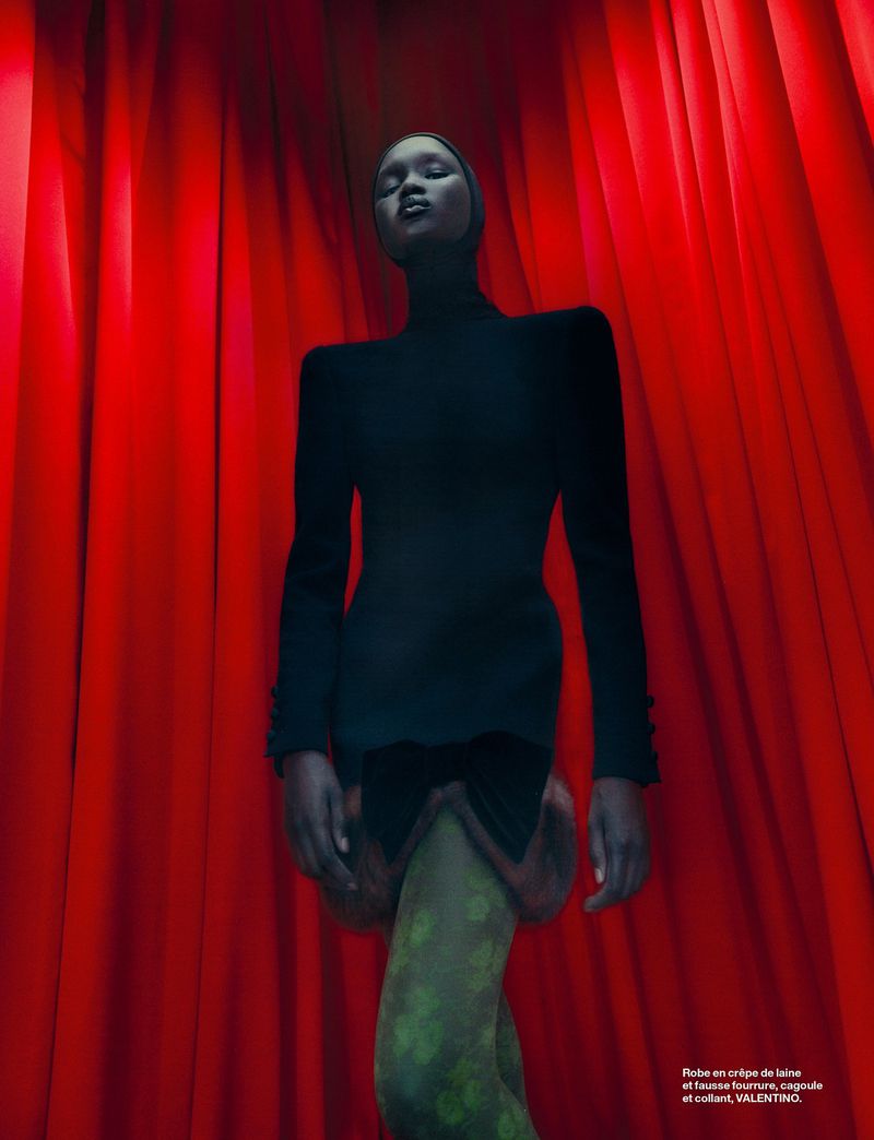 Fashion shot of a Black woman wearing a dark dress with floral tights, posing against a red curtain backdrop in a low-angle view.
