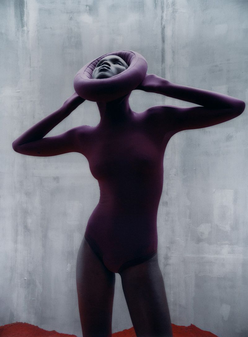 Fashion shot of a woman in a plum leotard and neck piece against a faded grey backdrop with a red accent at the bottom.