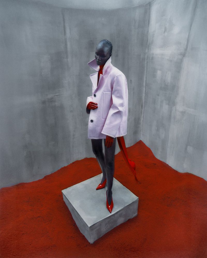 A dark-skinned model wearing a light pink jacket and red high heels poses on a concrete cube against a gray studio backdrop.