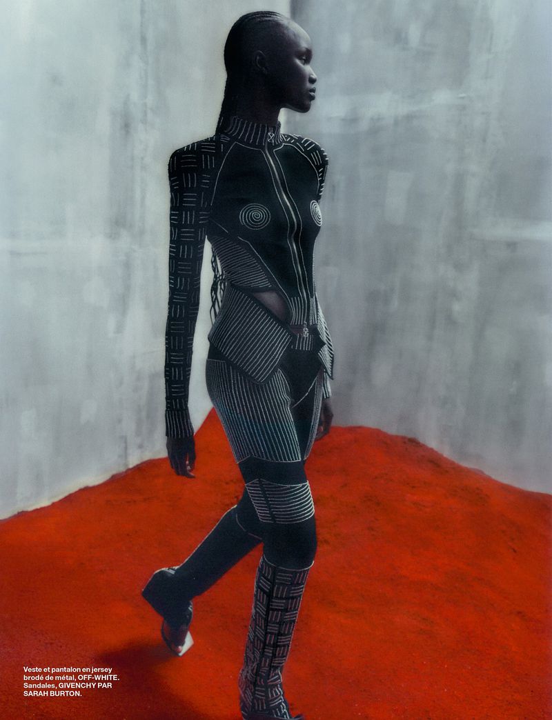 An African woman models patterned fashion in a studio, wearing a fitted jumpsuit with high boots, in a side view against a textured background.