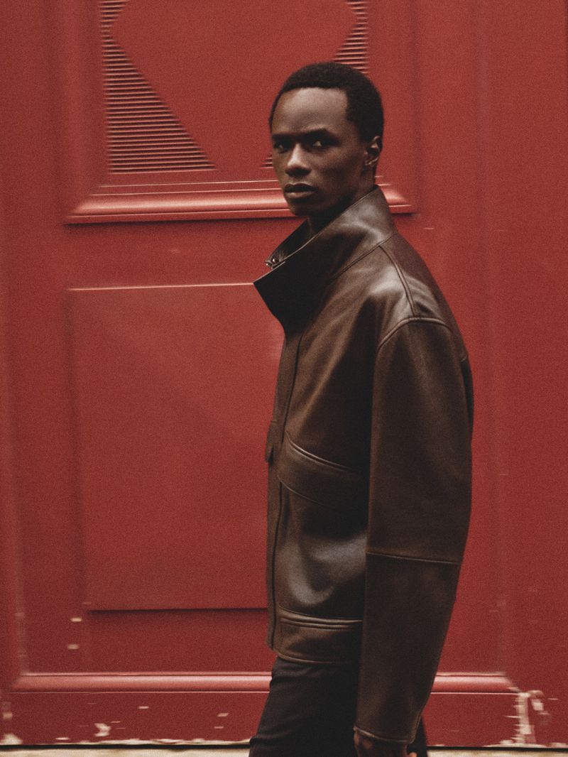 A young adult Black male stands in front of a textured red door, wearing a brown leather jacket, posing in an eye-level medium shot.