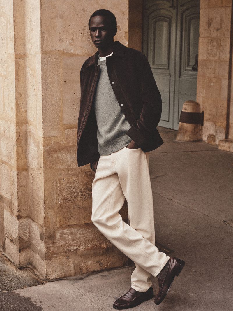 Adult male wearing dark jacket and white pants leans against stone wall near blue door on urban street, low angle perspective.
