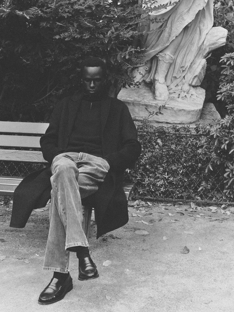 A man sits on a park bench in a black coat and jeans, with a statue and lush foliage in the background, captured in black and white.