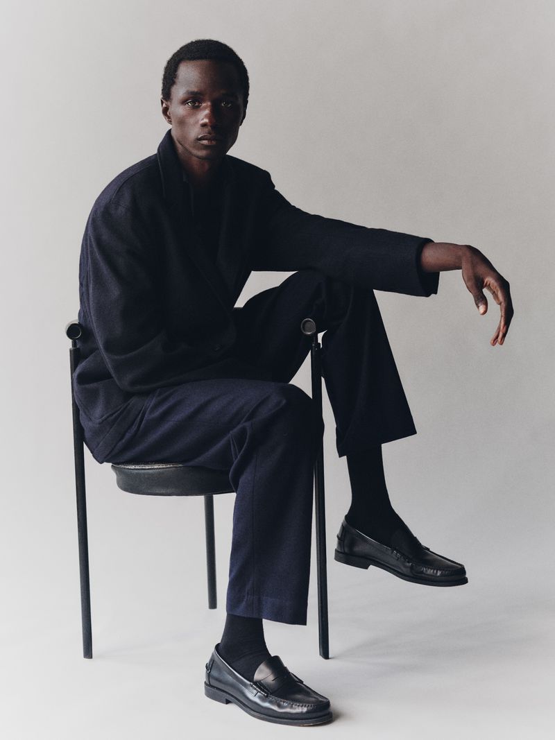 Adult male in dark workwear clothing and loafers, seated on a chair in a studio with a neutral background, portrait fashion shot.