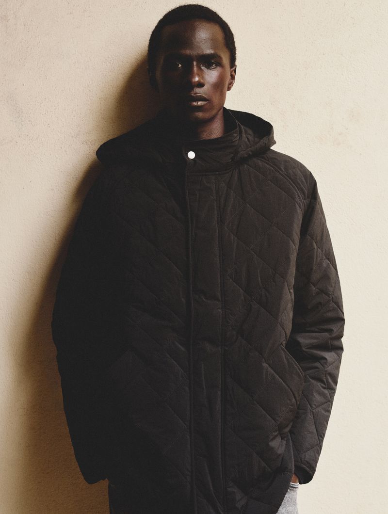 A dark-skinned male model poses in a hooded black quilted jacket against a neutral backdrop, highlighting fashion and studio photography.