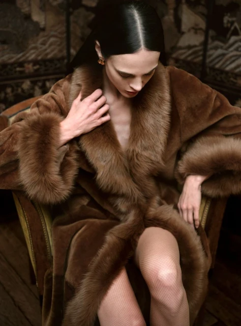 Elegant woman poses in a brown fur coat while seated in a chair, captured in a portrait-style shot with a dark background, fashion.