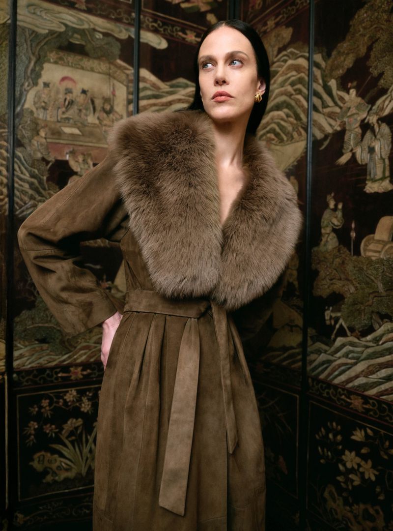 A woman in a brown coat with fur collar poses indoors against a dark decorative panel featuring paintings of landscapes and figures.