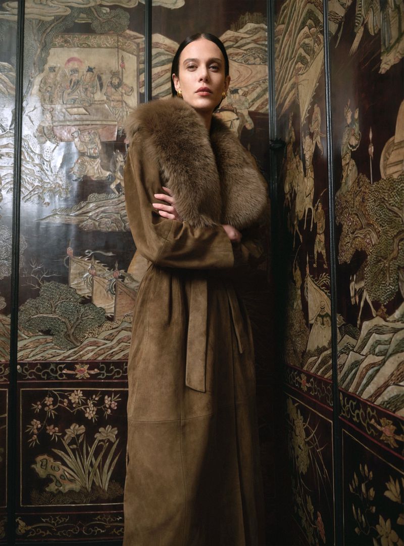 Elegant woman in long brown suede coat with fur collar, arms crossed, stands in front of ornate antique brown folding screen.