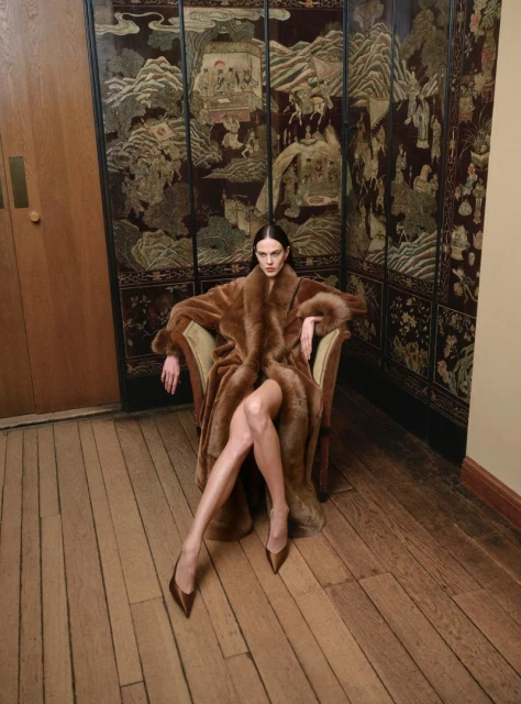Elegant woman in a fur coat sits in a chair in front of an ornate, antique folding screen on a wood floor wearing heels.
