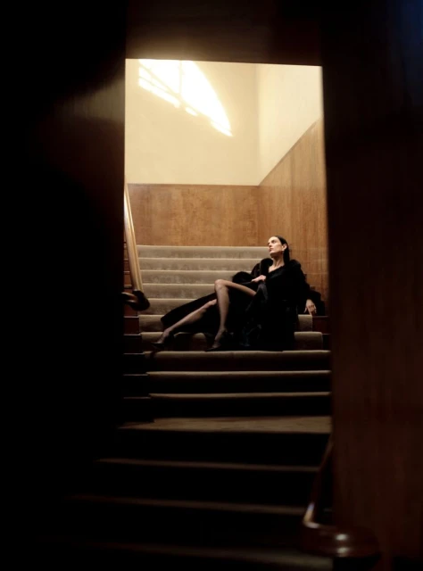 An adult female wearing a black dress and coat poses while sitting on carpeted stairs with a cream-colored wall and visible window light above.