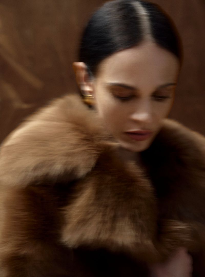 An adult woman with dark hair and tan skin wears a fur coat and gold earrings against a blurred brown background, portrait orientation.