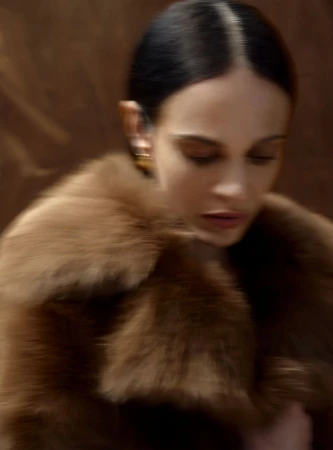 An adult woman with dark hair and tan skin wears a fur coat and gold earrings against a blurred brown background, portrait orientation.