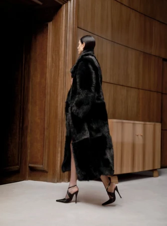 Woman in a black fur coat and high heels walking in a neutral-toned interior with wood paneling and a light floor. Fashion editorial shot.