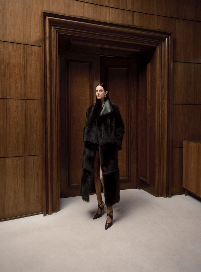 Fashion shot of a stylish adult woman in a long, dark coat posing in front of a wooden door indoors with neutral lighting and flooring.