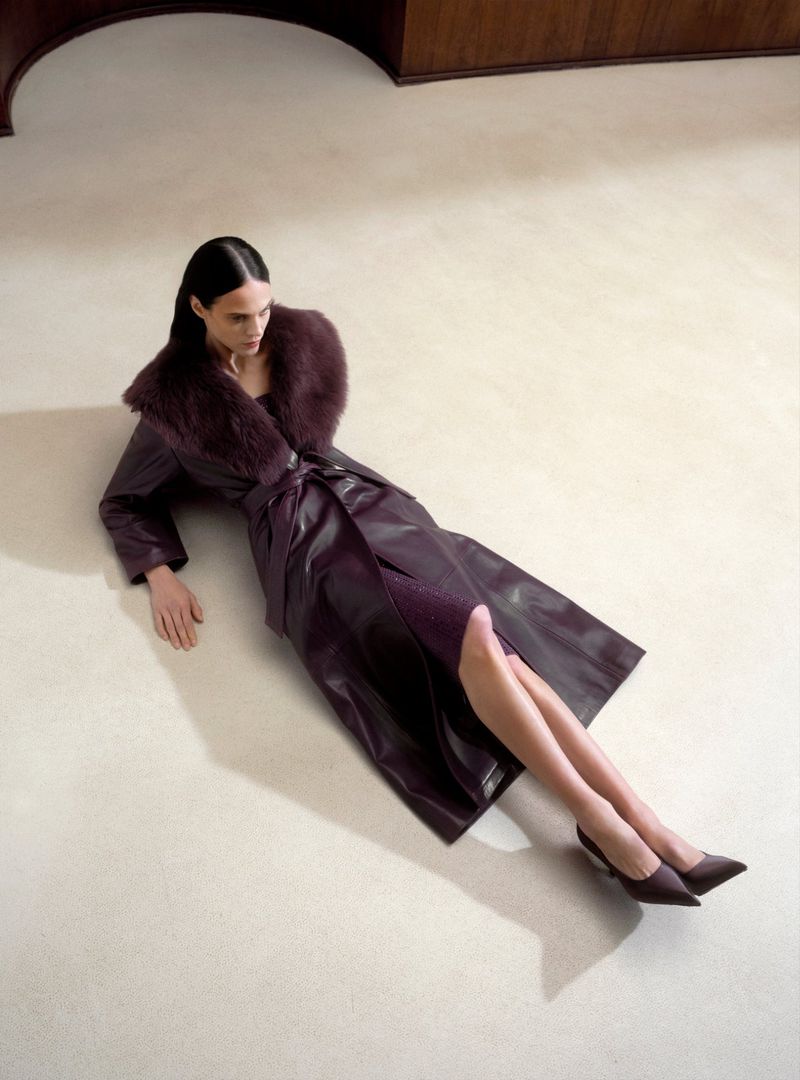 A woman in a plum leather coat with a fur collar poses on the floor, showcasing fashion and style in a studio setting with a neutral backdrop.