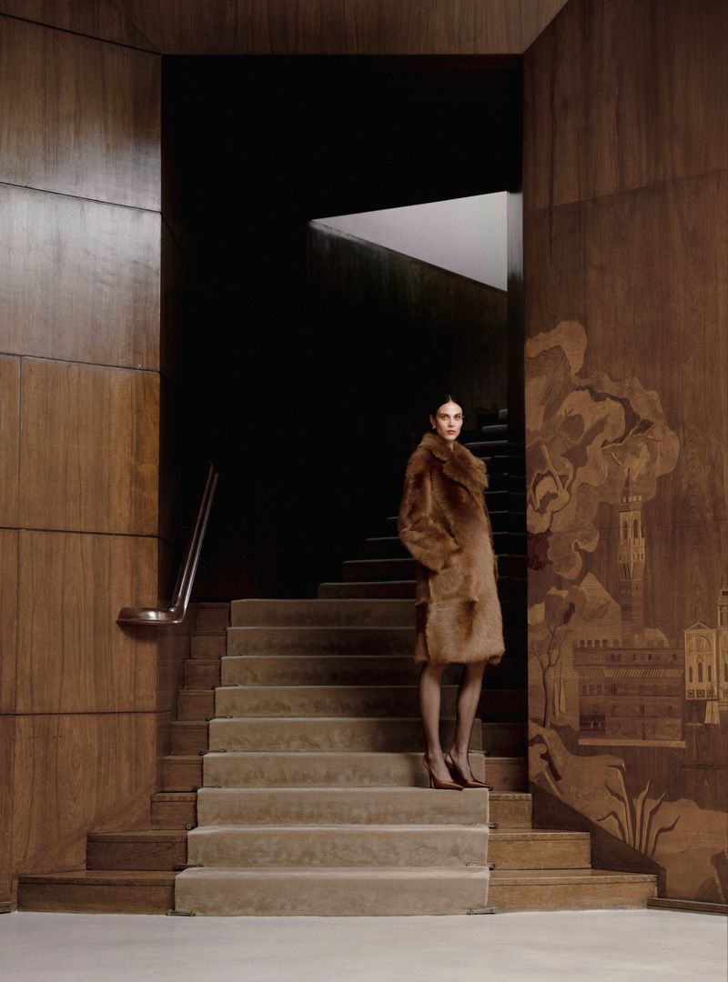 Woman poses on stairs in a brown fur coat with tall wood paneling. Editorial fashion photography in an indoor location with dark backdrop.