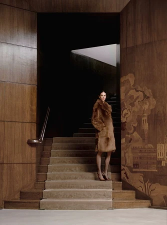 Woman poses on stairs in a brown fur coat with tall wood paneling. Editorial fashion photography in an indoor location with dark backdrop.