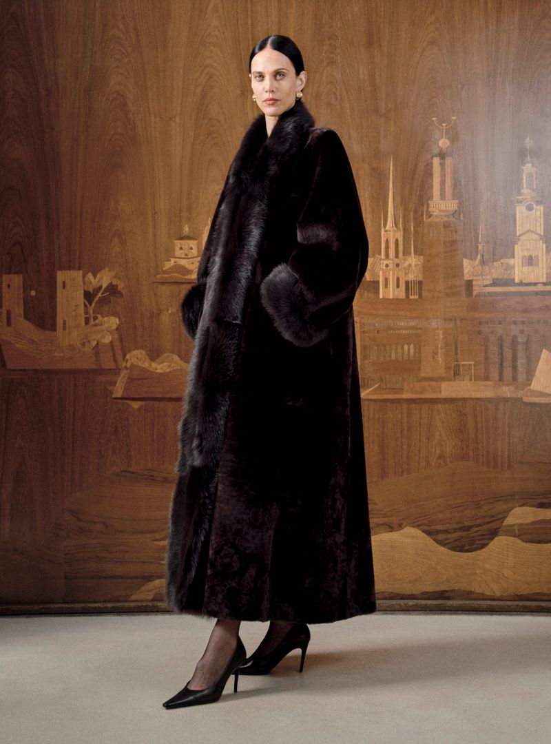 Elegant woman in long brown fur coat poses against wood-paneled backdrop. High fashion look with dark heels and gold earrings.