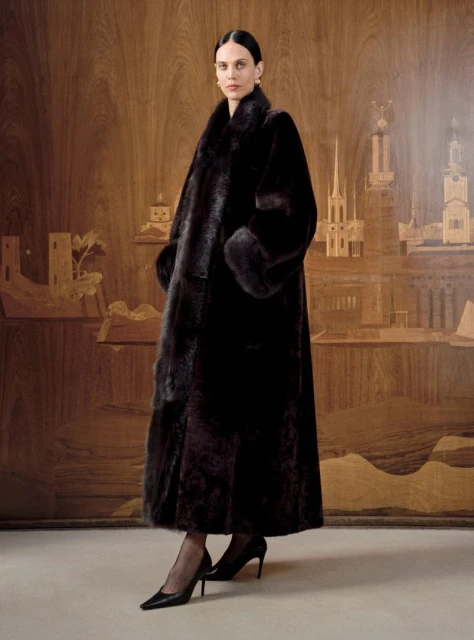 Elegant woman in long brown fur coat poses against wood-paneled backdrop. High fashion look with dark heels and gold earrings.