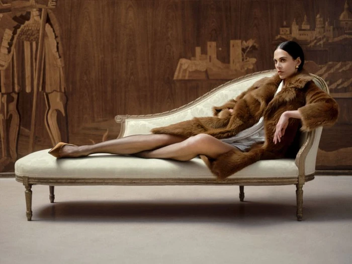 Model reclining on an antique chaise lounge wearing a brown fur coat against a wood-paneled wall.