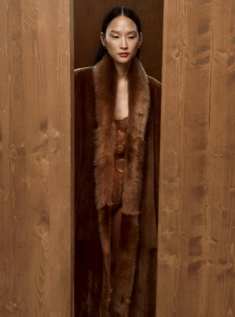 An Asian woman stands framed by a wooden structure, wearing a brown faux fur coat and dress, in a warmly lit indoor setting.