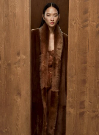 An Asian woman stands framed by a wooden structure, wearing a brown faux fur coat and dress, in a warmly lit indoor setting.