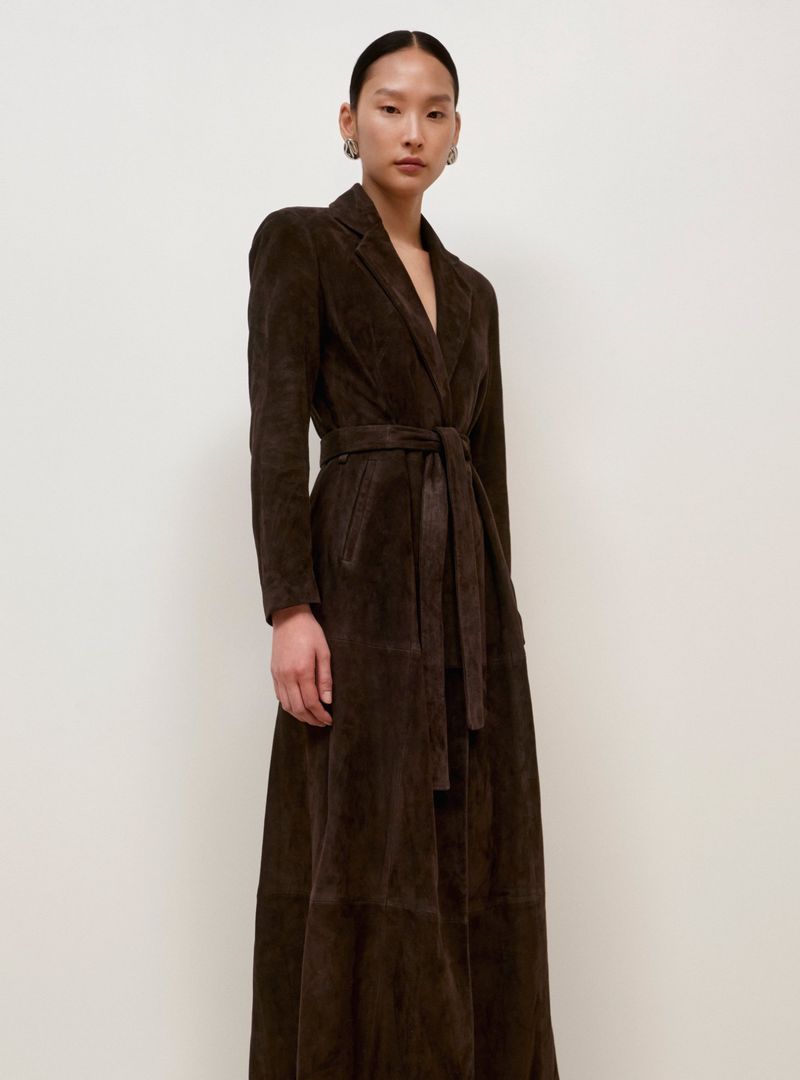 A woman models a long-sleeved, ankle-length brown coat with a matching tie belt indoors against a neutral-tone wall.