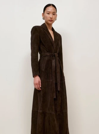 A woman models a long-sleeved, ankle-length brown coat with a matching tie belt indoors against a neutral-tone wall.