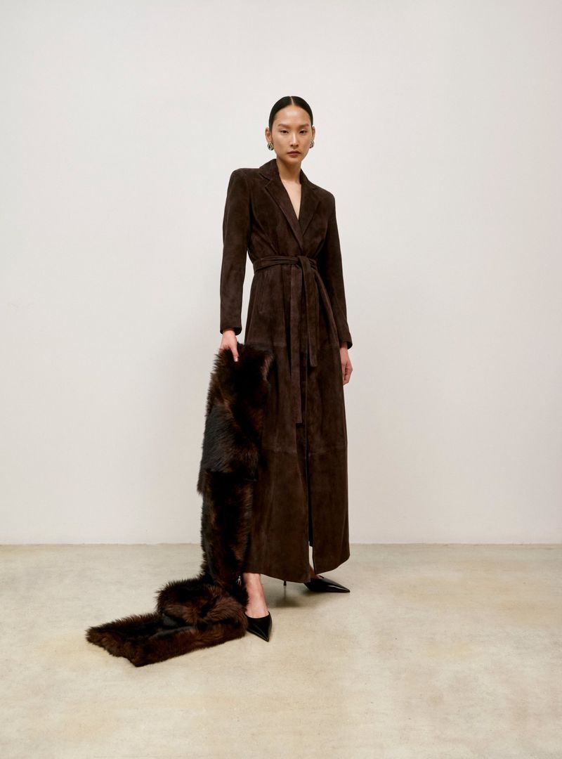 A slender Asian woman in a long belted coat poses indoors, holding a fur stole that trails onto a concrete floor, under bright lighting.