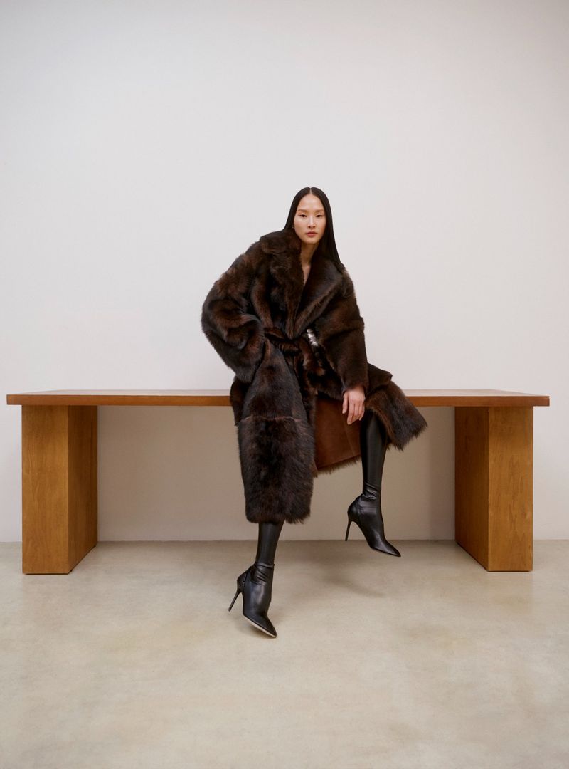 A woman in a dark brown fur coat and black boots sits on a wooden table against a white wall in a studio environment.