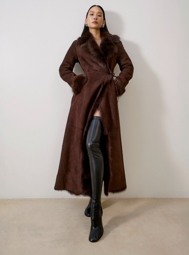A stylish woman in a brown fur-trimmed coat and black thigh-high boots poses in a studio against a neutral backdrop, showcasing a fashion-forward look.