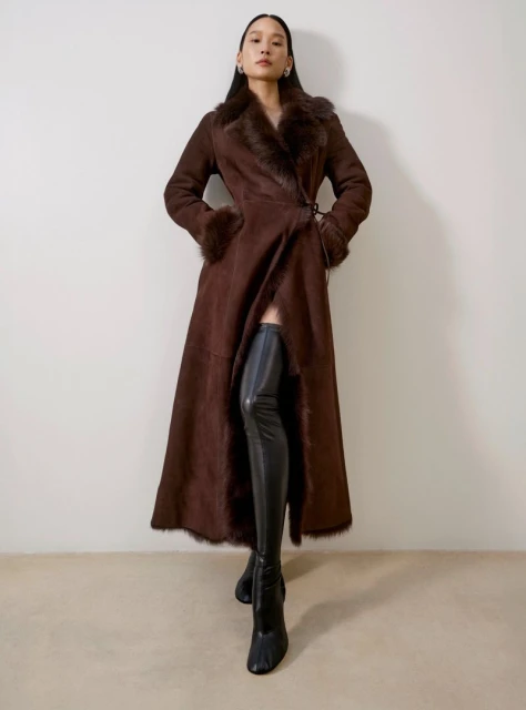 A stylish woman in a brown fur-trimmed coat and black thigh-high boots poses in a studio against a neutral backdrop, showcasing a fashion-forward look.