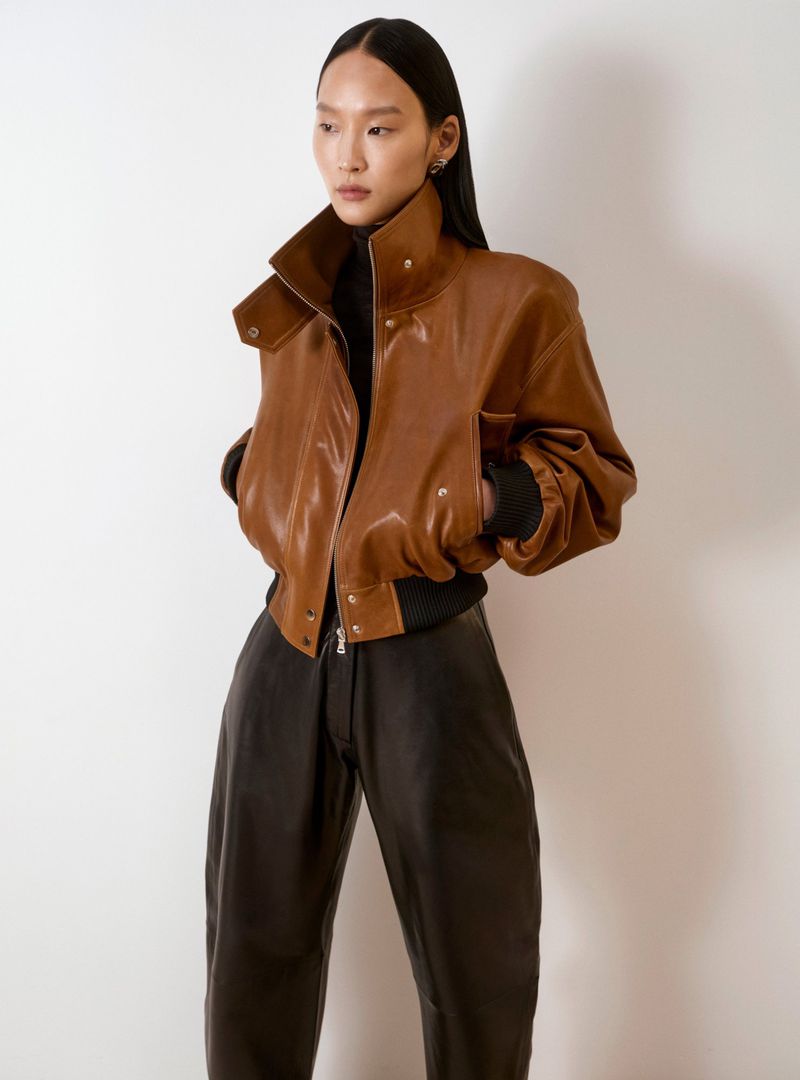 Asian woman wearing a brown leather bomber jacket and black pants poses against a neutral background in a fashion studio shot.