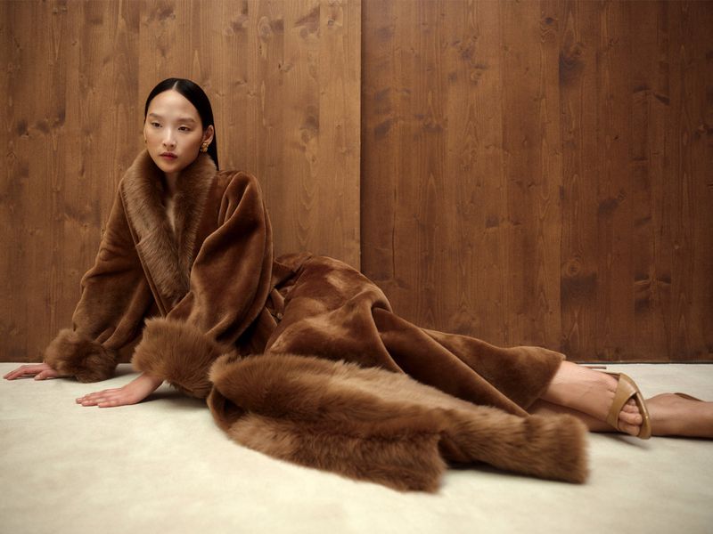 An Asian female model poses in a luxurious brown faux-fur coat and nude heels against a warm-toned wood paneled background indoors.