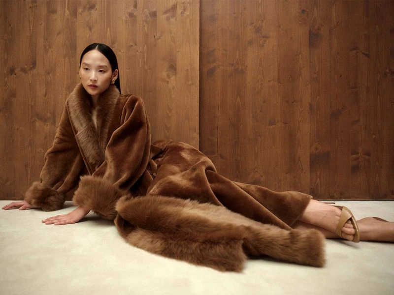 An Asian female model poses in a luxurious brown faux-fur coat and nude heels against a warm-toned wood paneled background indoors.