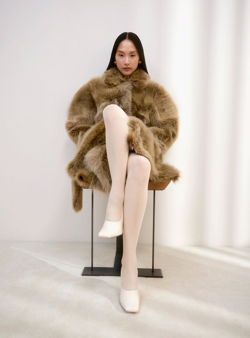 An Asian woman sits on a stool wearing a brown faux fur coat, white tights, and white slip-on shoes in a brightly lit photo studio.