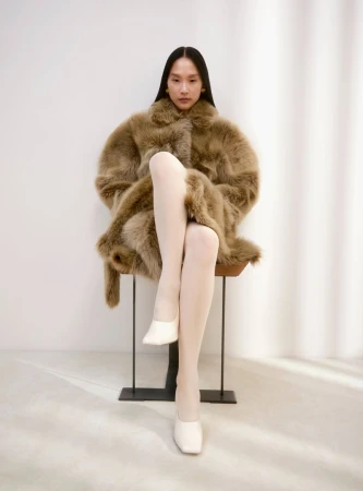 An Asian woman sits on a stool wearing a brown faux fur coat, white tights, and white slip-on shoes in a brightly lit photo studio.