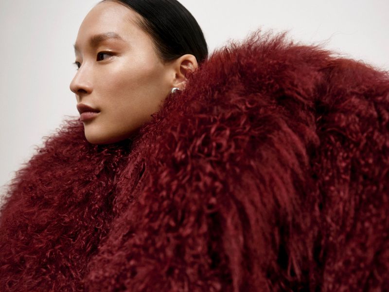 Asian woman with clean makeup and slicked-back hair wearing a burgundy, furry coat posing against a white background.