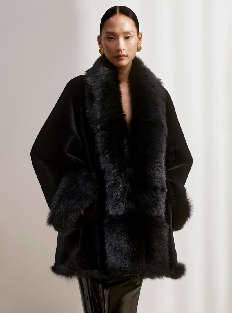 Portrait of an adult woman wearing a black faux fur coat and gold earrings, posing in an indoor photo studio with a neutral background.