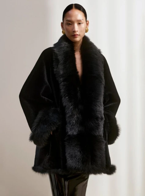 Portrait of an adult woman wearing a black faux fur coat and gold earrings, posing in an indoor photo studio with a neutral background.