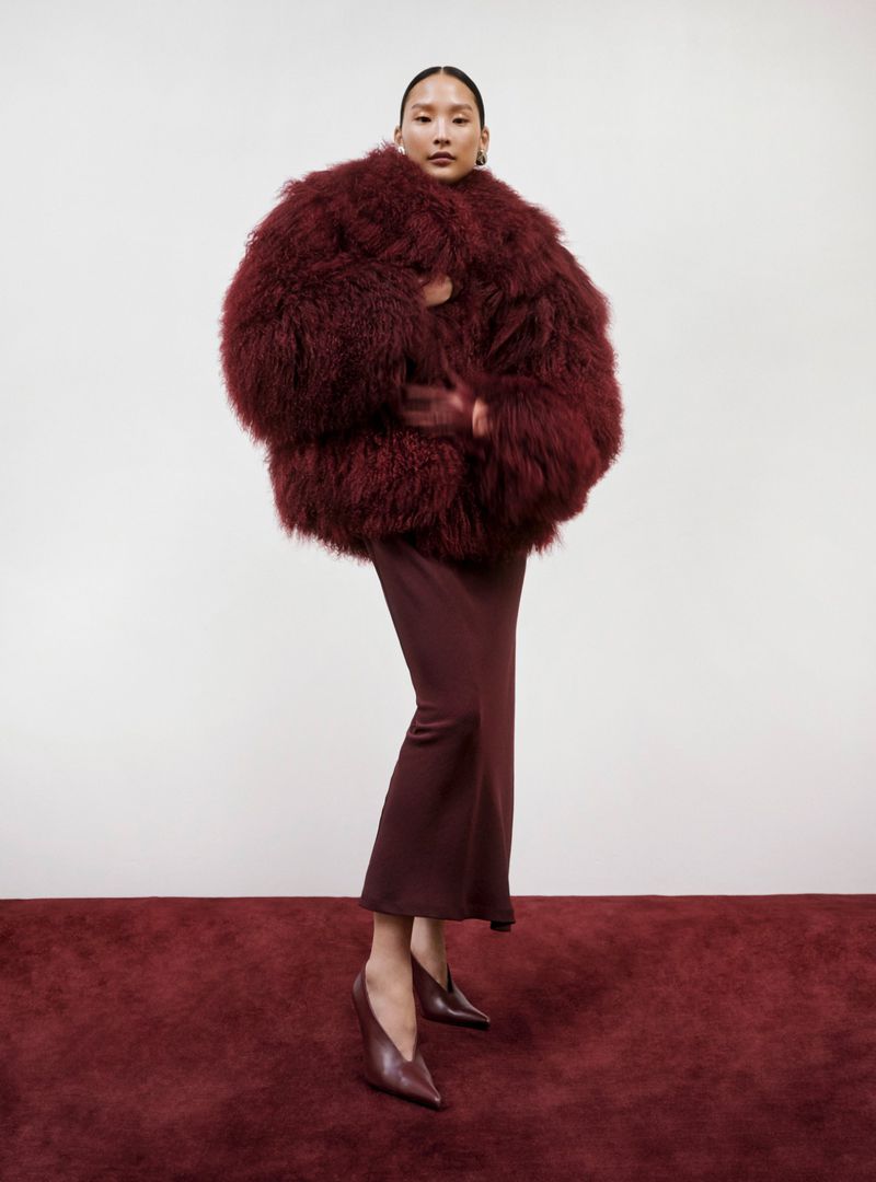 A woman with dark hair poses indoors wearing a large, fluffy burgundy coat, a dark burgundy dress, and dark burgundy heels on a red carpet.
