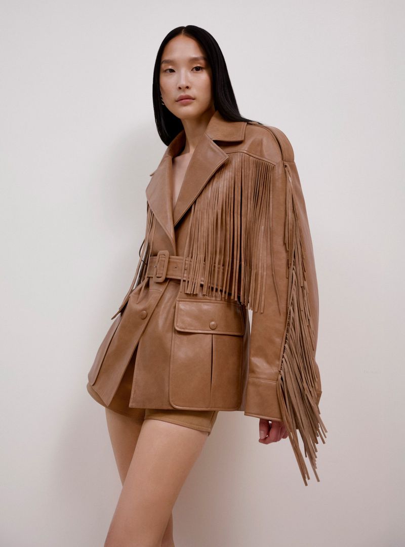 Portrait of an Asian woman wearing a tan leather jacket with fringe details, posing in front of an off-white background. Fashion shot.