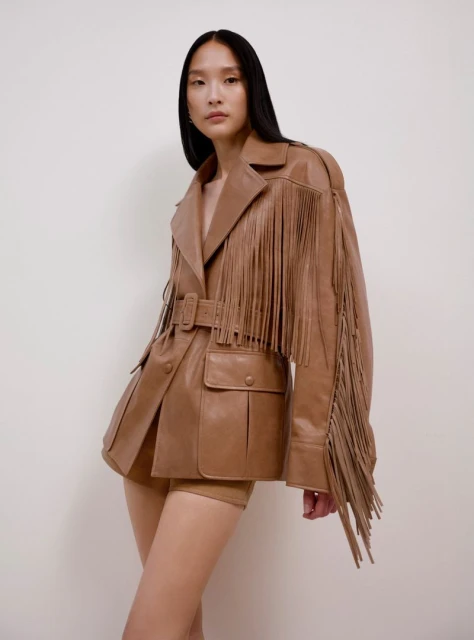 Portrait of an Asian woman wearing a tan leather jacket with fringe details, posing in front of an off-white background. Fashion shot.